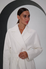 Woman wearing a white coat and black pants, holding a black handbag, against a plain background.