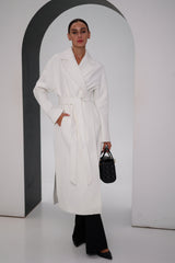 Woman wearing a white coat and black pants, holding a black handbag, against a plain background.