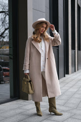 Woman in a long beige coat and hat standing on a city street.