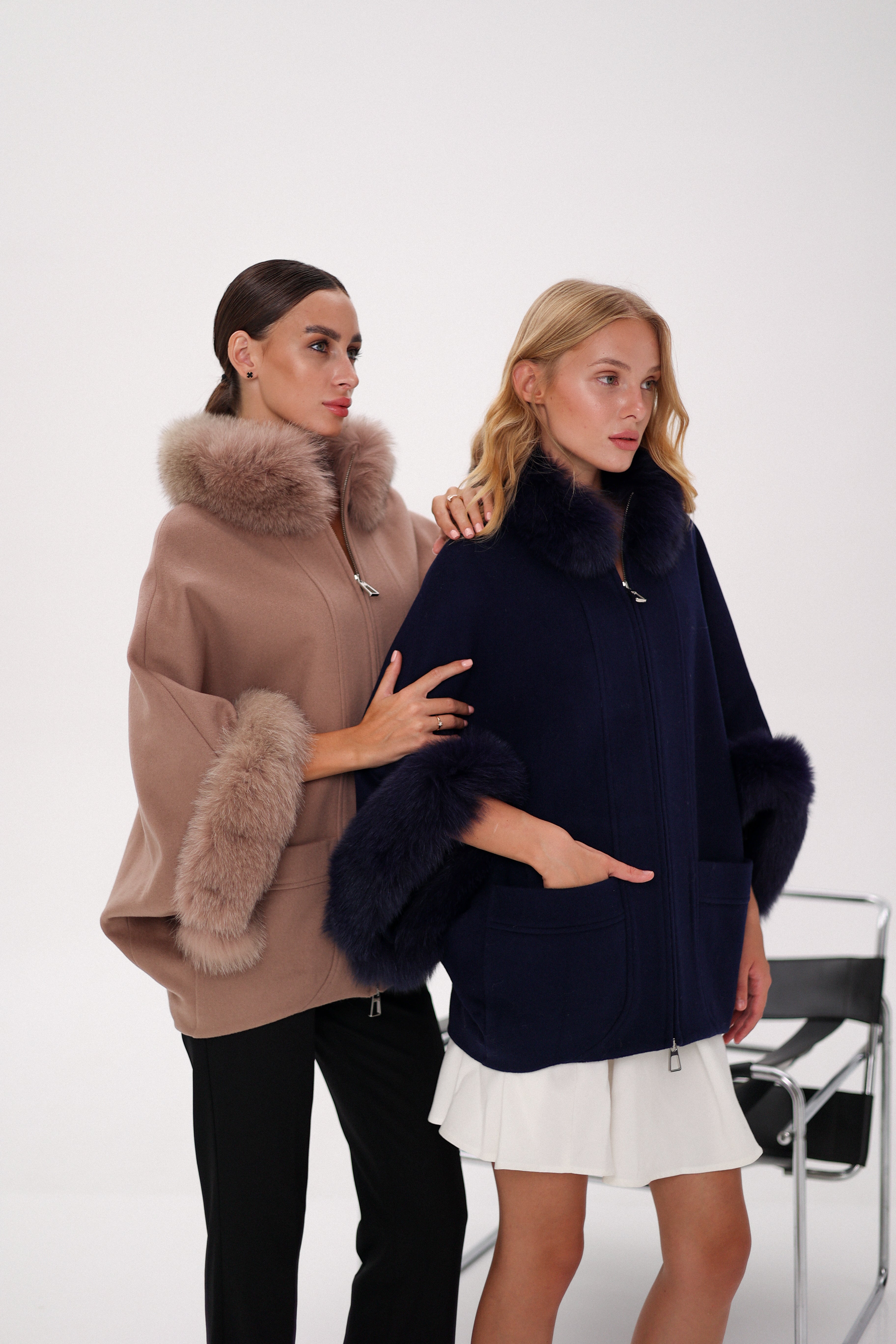 Two women wearing fur-trimmed ponchos against a white background