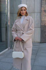 Woman in a beige coat and white hat holding a white handbag against a gray building background