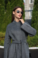 Woman wearing a gray coat and sunglasses outdoors with greenery in the background
