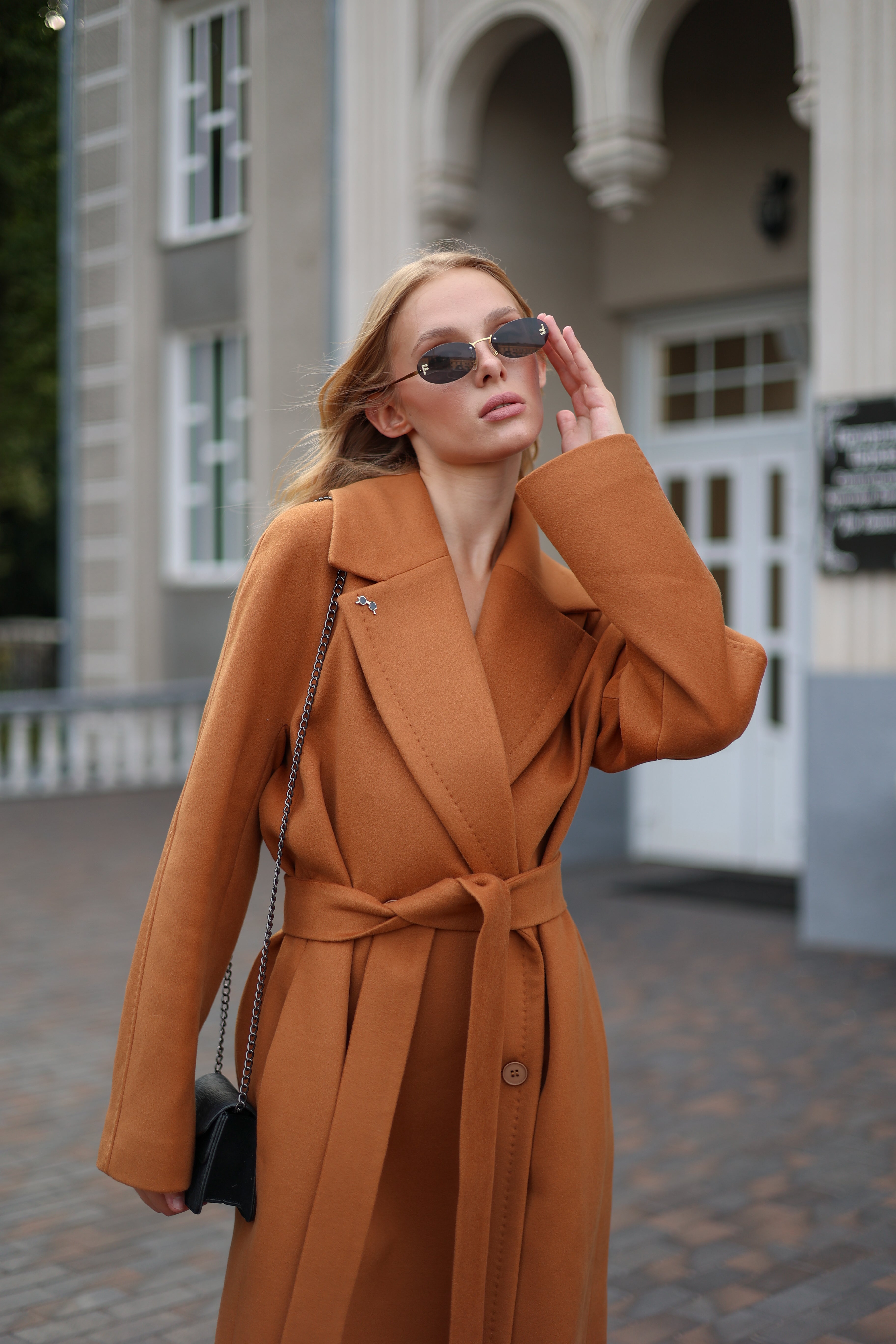 Woman in a brown coat standing outdoors with a building in the background