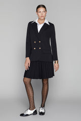 Woman wearing a black blazer with gold buttons and a pleated skirt on a gray background