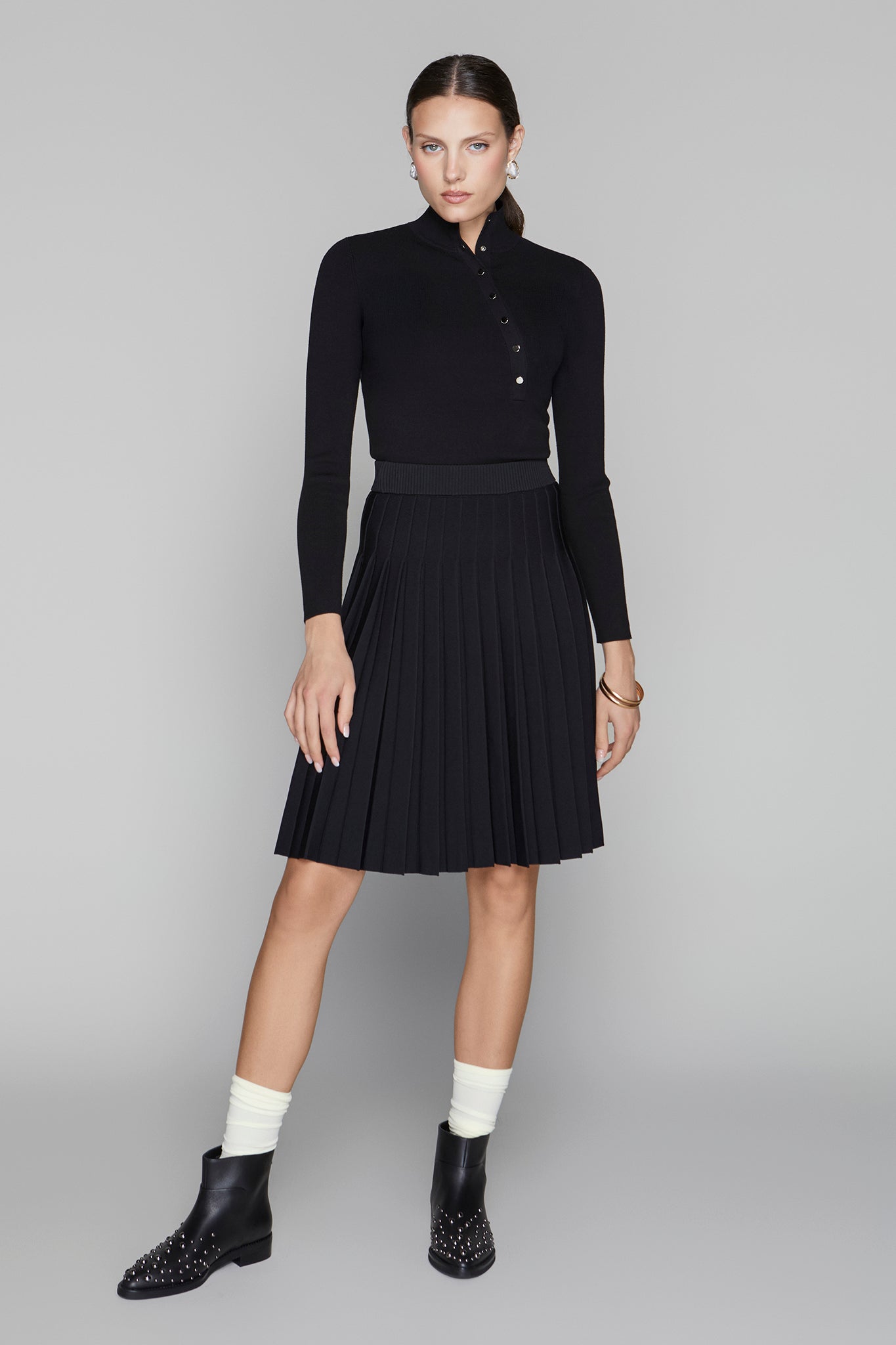 Woman wearing a black outfit with a high collar and pleated skirt on a gray background