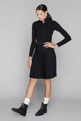 Woman wearing a black long-sleeve top and pleated skirt with black boots on a gray background