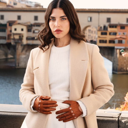 Woman in a beige coat and white shirt with brown gloves standing by a river with buildings in the background