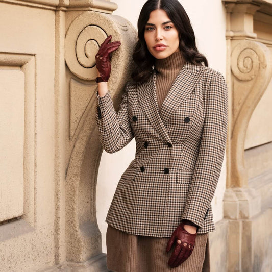 Woman in a checkered blazer and brown gloves standing against a decorative wall.