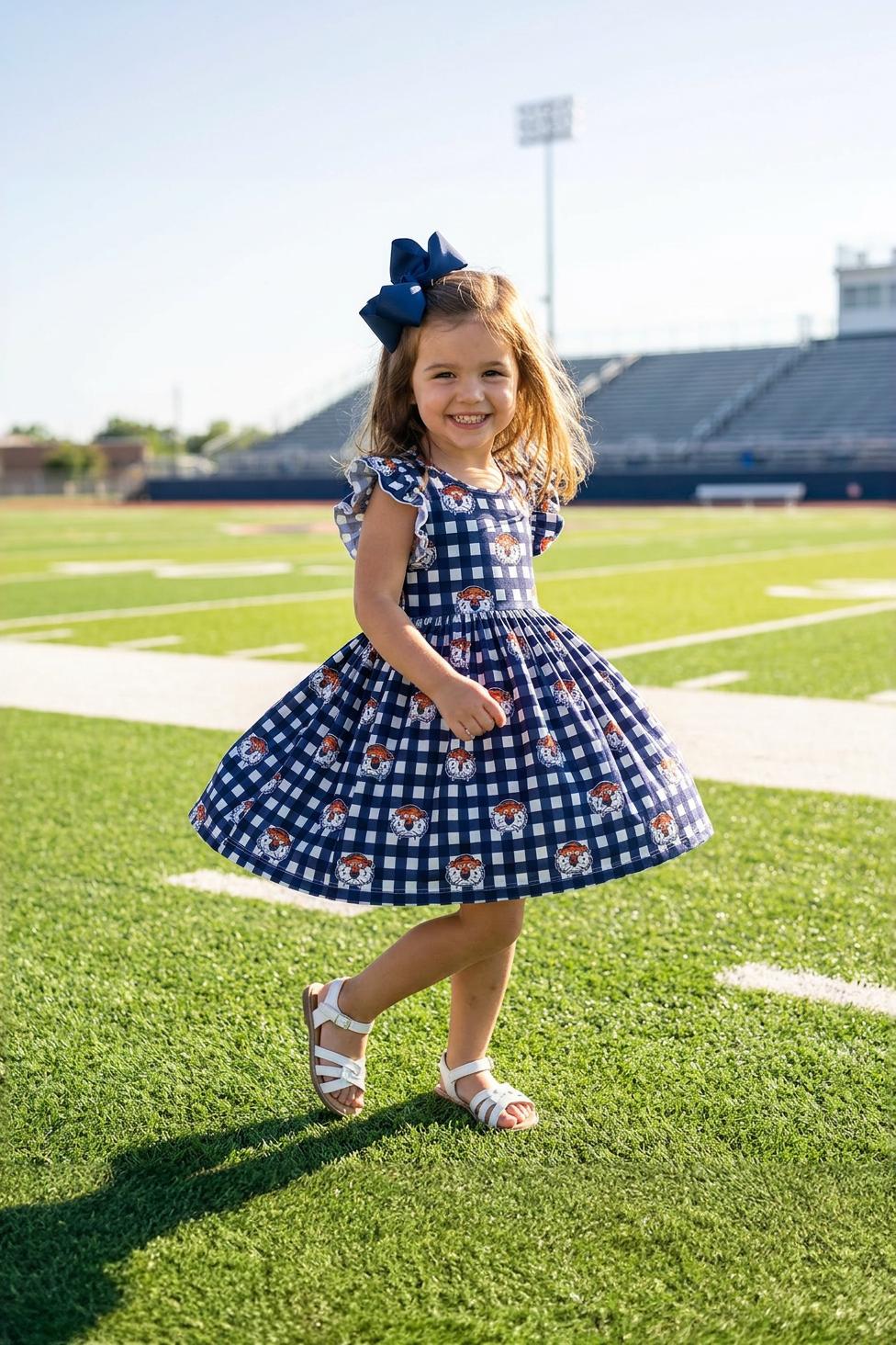 Young girl in a blue checkered dress with a bow standing on a football field.