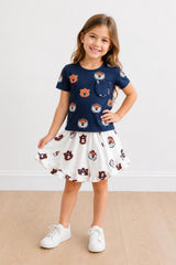 Young girl wearing a navy blue top with a pocket and a white skirt with a pattern, standing on a beige floor.