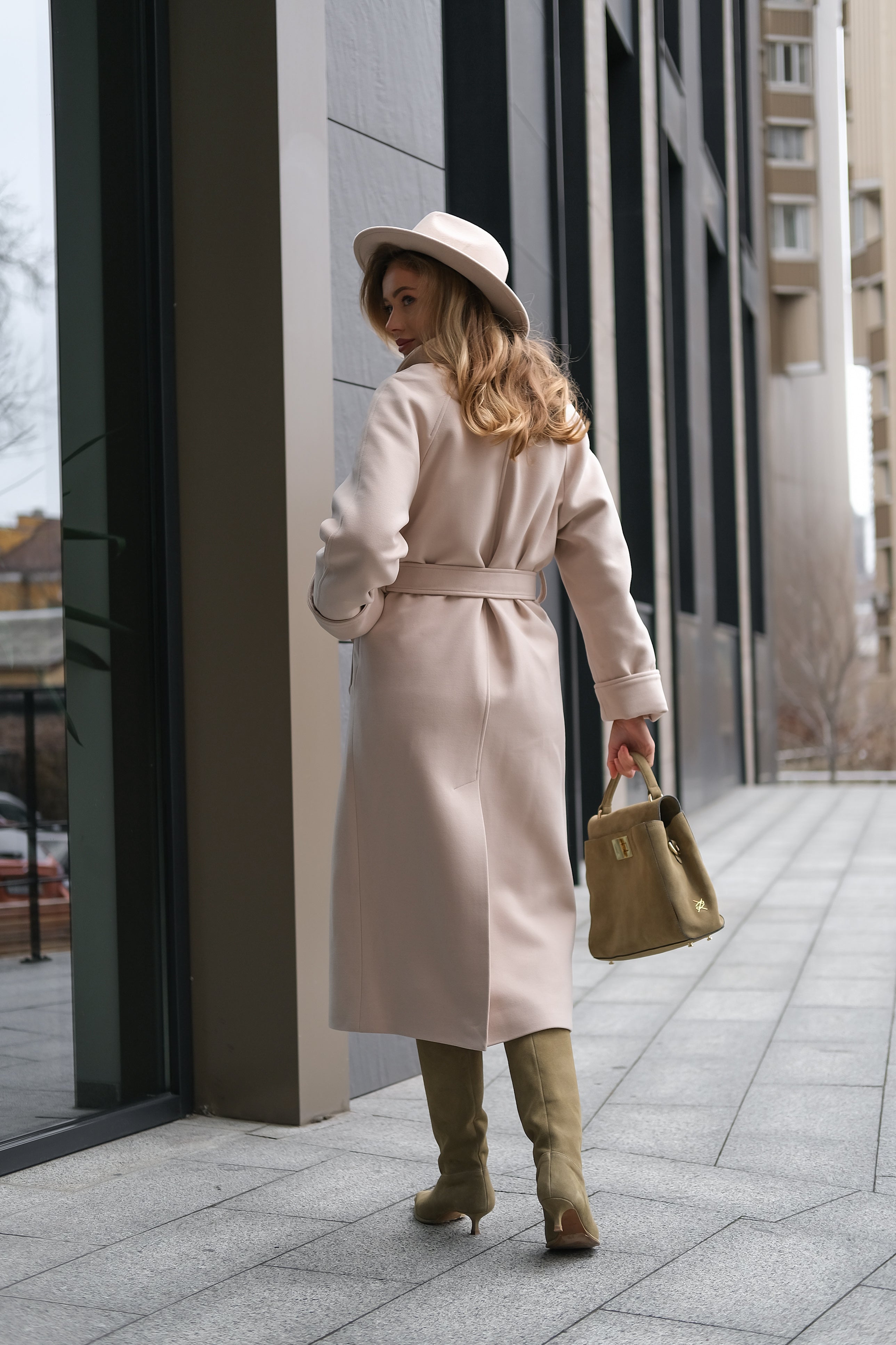 Woman in a long coat and hat walking on a city sidewalk.