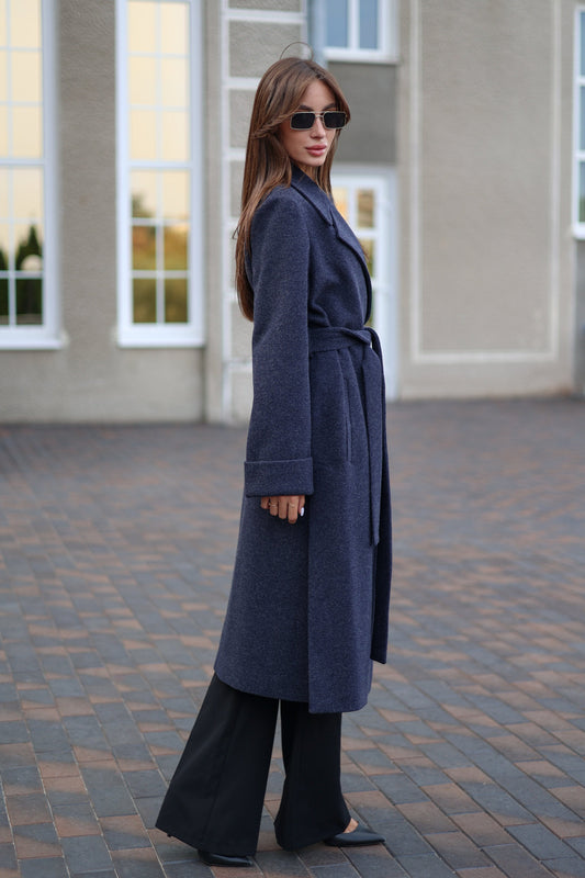 Woman wearing a long blue coat standing on a paved area with a building in the background