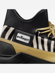 Black and gold sneaker with zebra pattern and 'BYMANYC' label on a white background