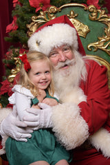 A smiling young girl in a green dress and white gloves hugs Santa Claus, who wears his red suit and white beard while seated in a green and gold festive chair.
