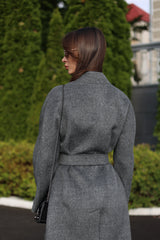 Woman wearing a gray coat walking outdoors with greenery in the background