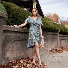Woman in a floral dress standing outdoors near a stone wall with greenery and fallen leaves.