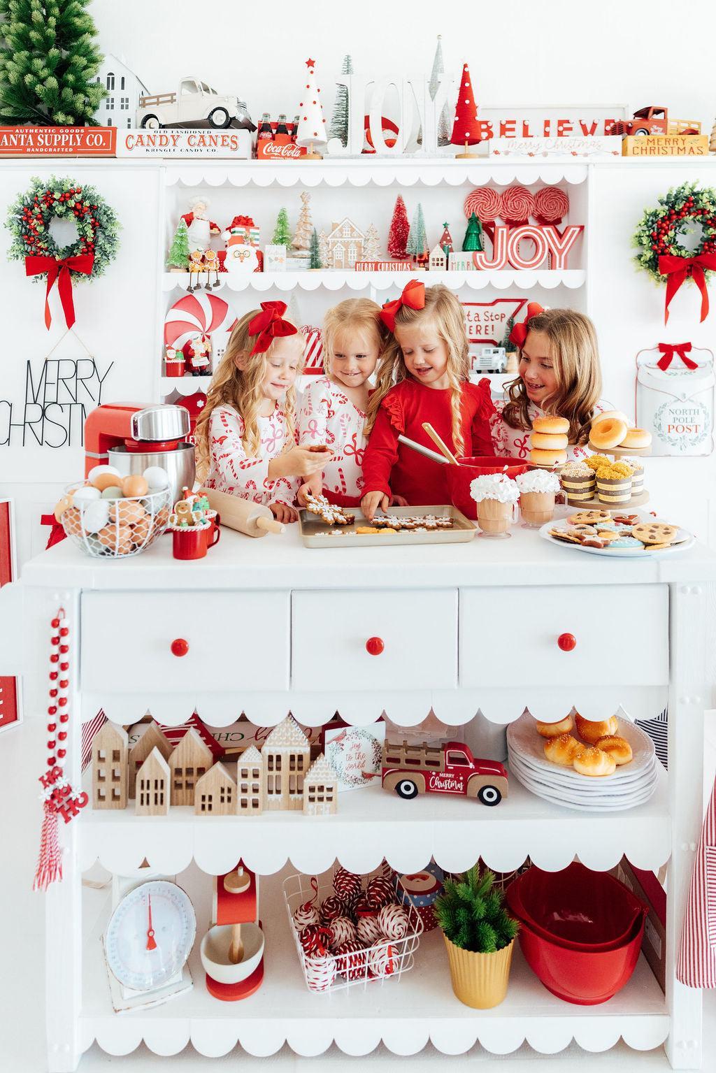 A festive holiday kitchen scene with three young girls standing behind a white scalloped counter filled with cookies, pastries, red bowls, and Christmas decorations. The shelves above are decorated with candy canes, miniature trees, signs reading 'JOY' and 'MERRY CHRISTMAS', and red wreaths.