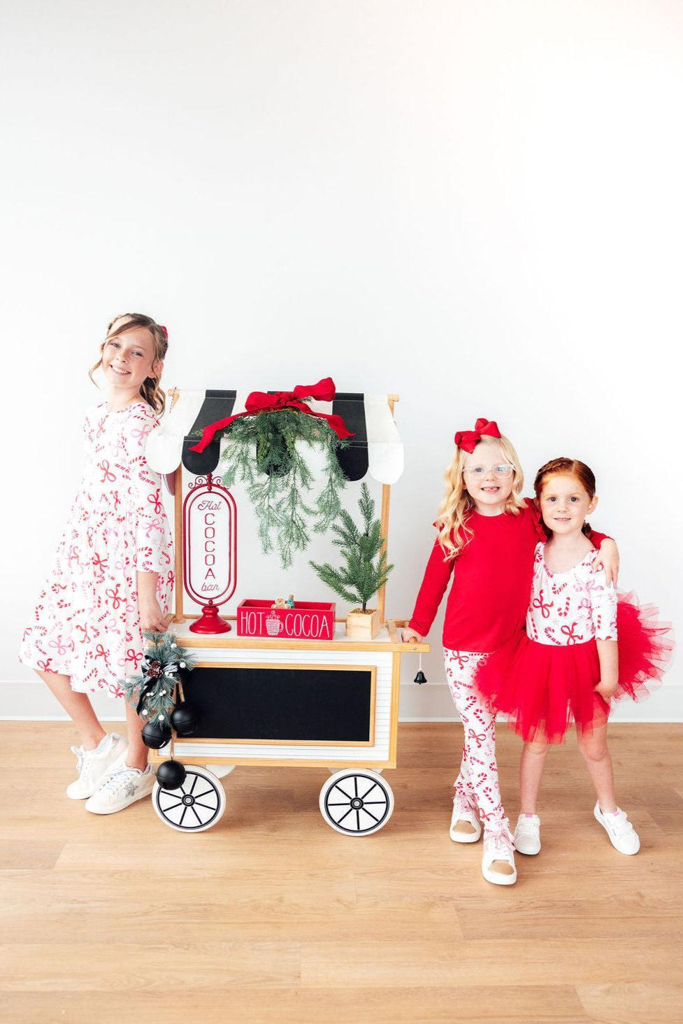 Three young girls in festive holiday outfits pose next to a decorative wooden toy popcorn cart with green pine garland and red accents, set against a plain white wall and light wood floor.