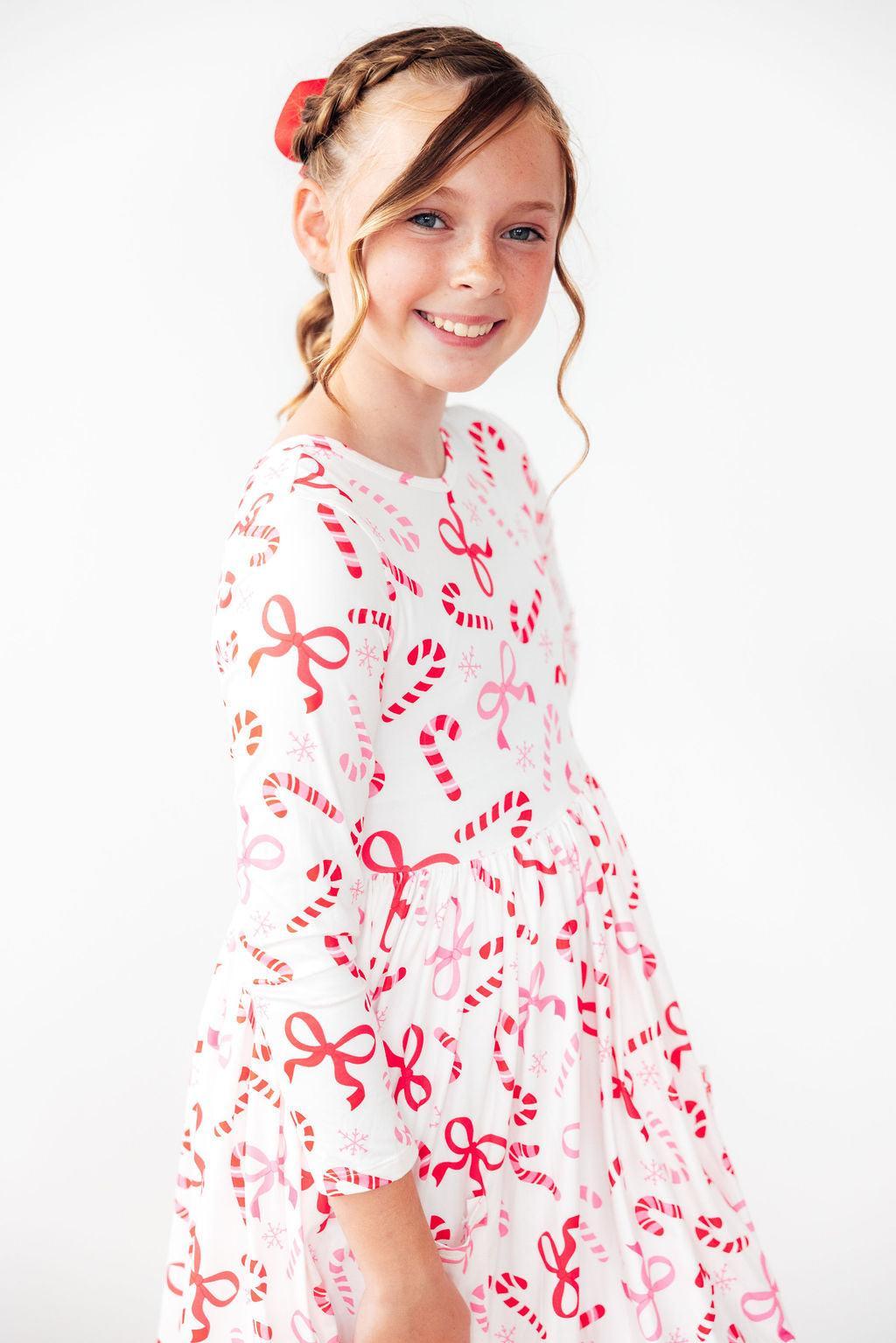 A young girl with a braided ponytail and a red hair bow is wearing a white long-sleeve dress with a red and pink candy cane and bow pattern, photographed against a clean white background.