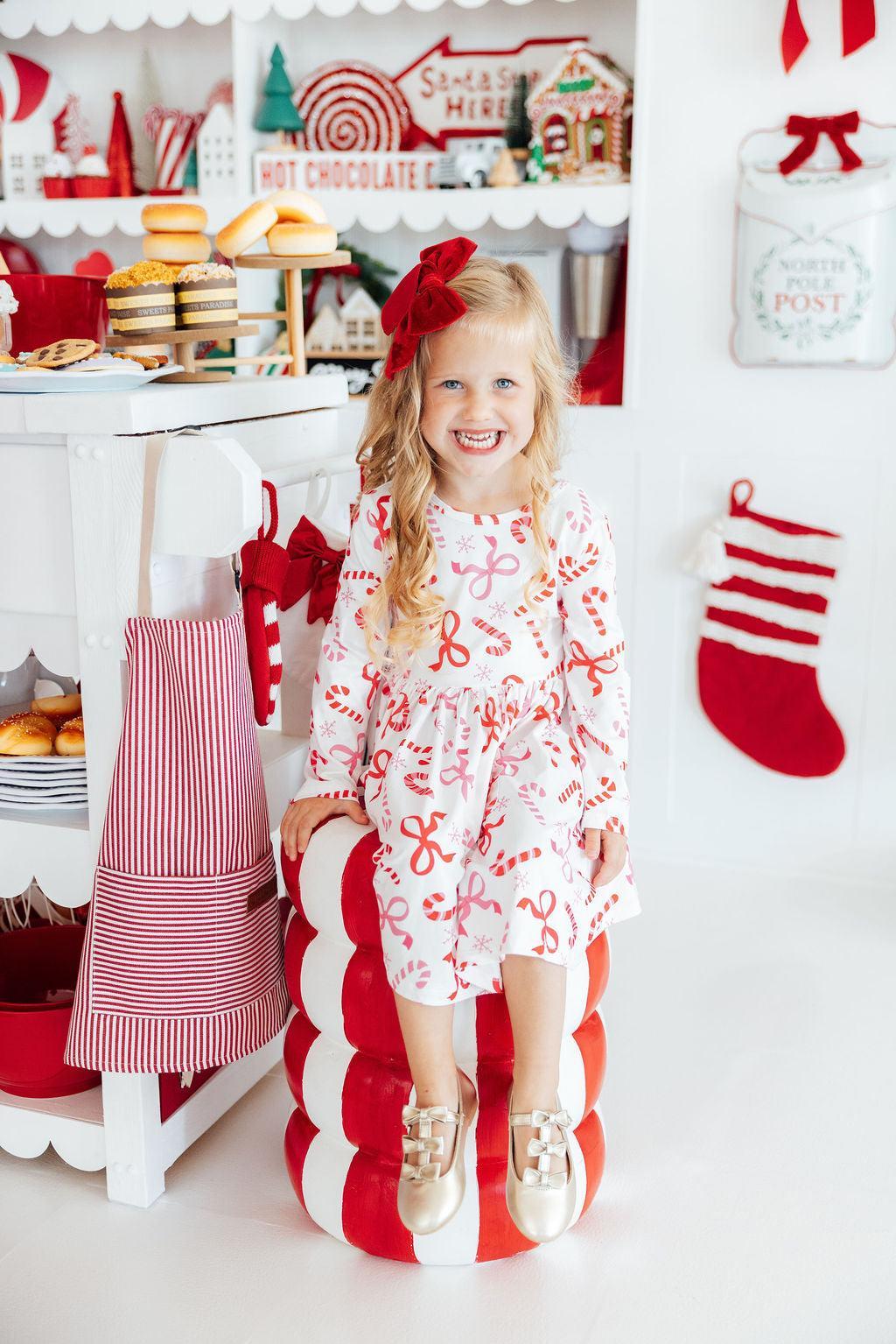 A young girl with blonde hair and a red bow sits on a red and white striped candy cane-shaped pouf in a festive holiday kitchen. She is wearing a white long-sleeve dress decorated with red and pink candy cane and bow patterns, paired with gold shoes.