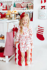 A young girl with blonde hair and a red bow sits on a red and white striped candy cane-shaped pouf in a festive holiday kitchen. She is wearing a white long-sleeve dress decorated with red and pink candy cane and bow patterns, paired with gold shoes.