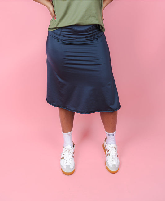 Person wearing a navy skirt, green top, white sneakers, and socks on a pink background