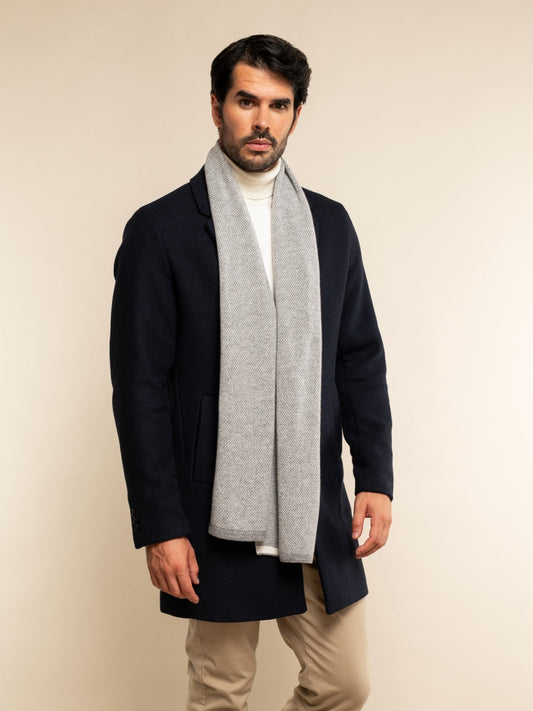 Man wearing a black coat, white turtleneck, beige pants, and a gray scarf on a beige background