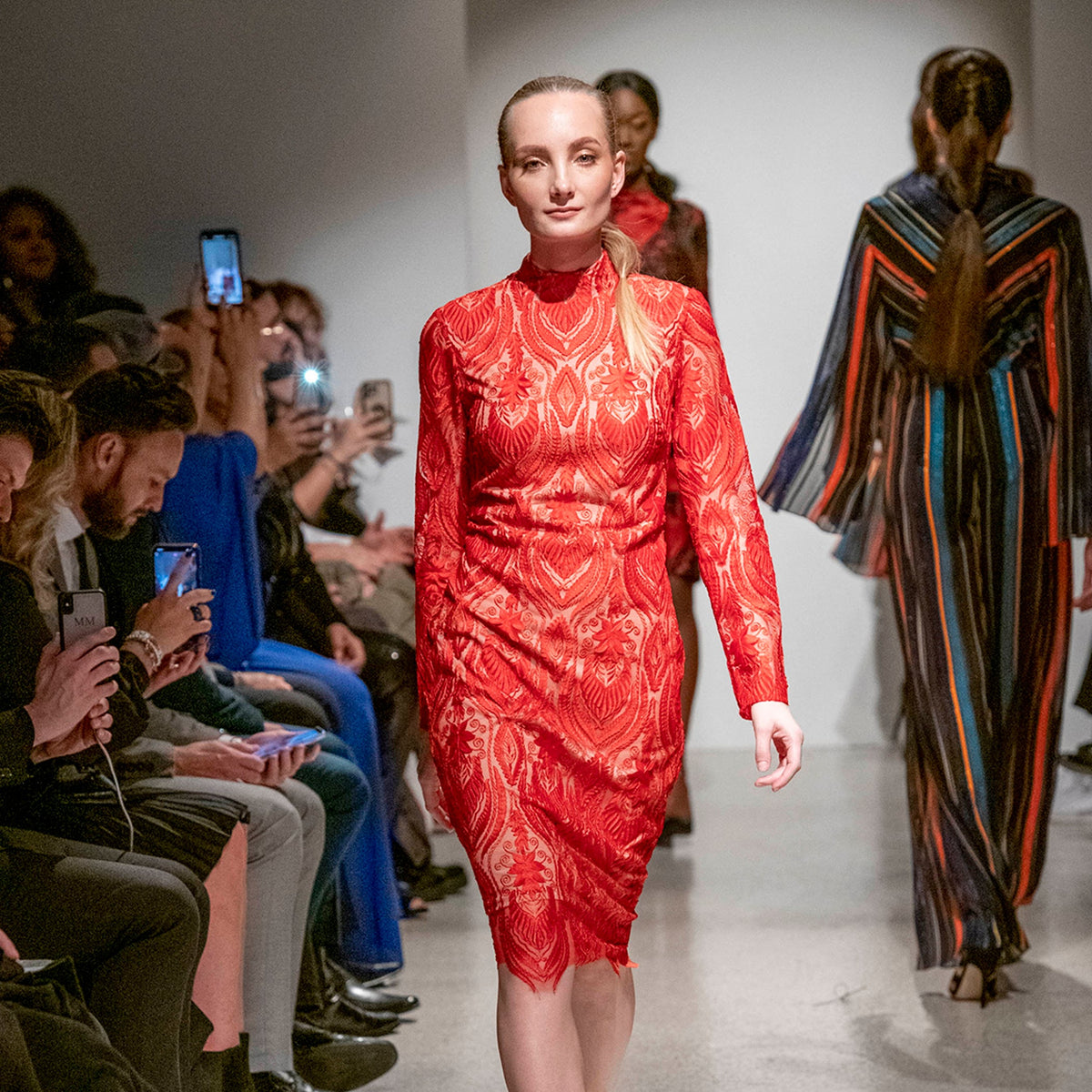 Model in a red dress walking on a fashion runway with an audience taking photos.