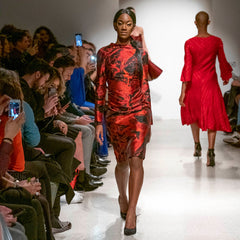Model walking down a runway wearing a red and black dress, with audience members taking photos.