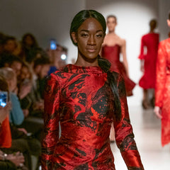 Model wearing a red and black patterned dress on a fashion runway with audience and other models in the background.