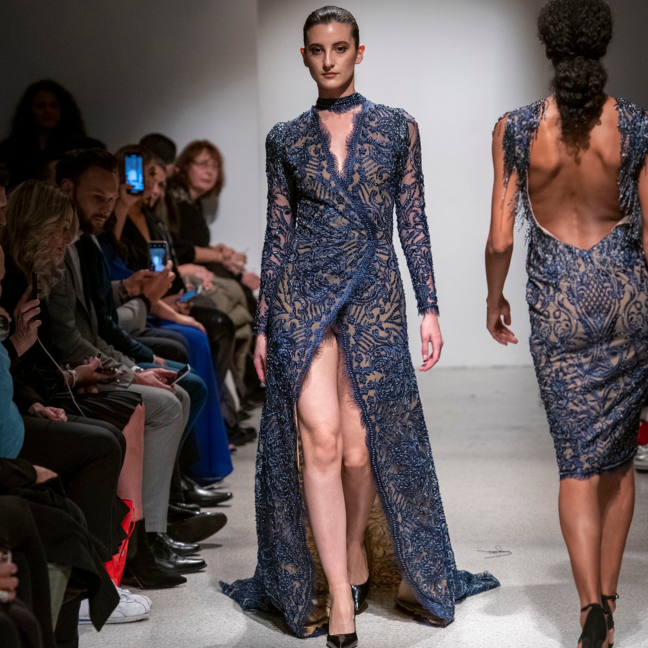 Model wearing a blue lace dress on a fashion runway with an audience.
