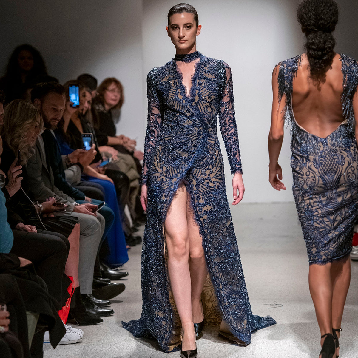 Model wearing a blue lace dress on a fashion runway with an audience.