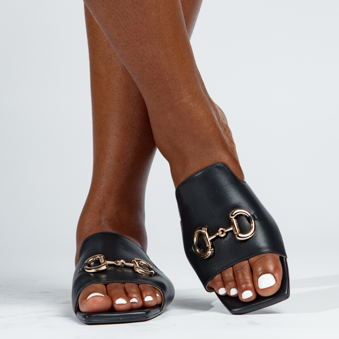 Black sandals with gold hardware worn by a person on a white background