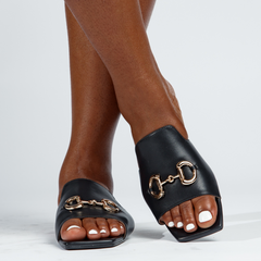 Black sandals with gold hardware worn by a person on a white background
