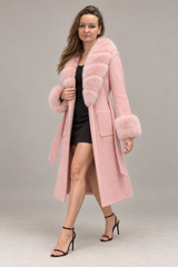 A woman modeling a long light pink coat with fluffy faux-fur trim at the collar, cuffs, and hem, worn over a black dress and black high-heeled sandals against a plain gray studio background.
