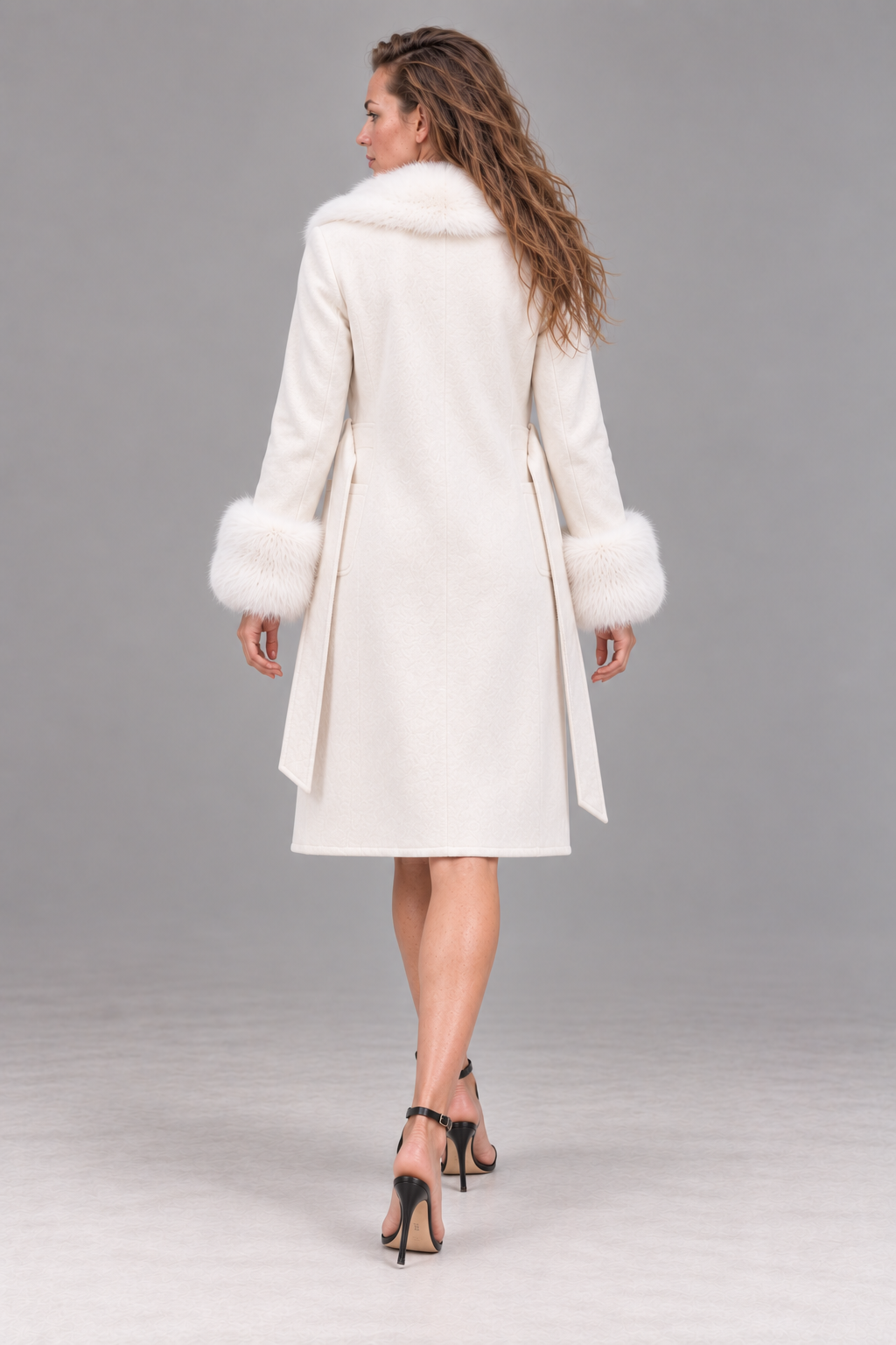 A woman shown from the back wearing a long cream-colored coat with fluffy fur trim at the collar and cuffs, paired with black high-heeled sandals, against a neutral gray studio background.