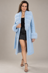 A woman modeling a light blue faux fur coat with a matching collar and cuffs, worn over a black top and black leather skirt, against a solid beige studio background.