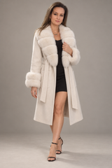 A woman modeling a long cream-colored faux fur coat with a matching belt, worn over a black dress and black high-heeled sandals, against a neutral gray studio background.