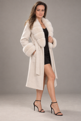 A woman modeling a long cream-colored coat with a fluffy faux-fur collar and cuffs, worn over a black dress and black high-heeled sandals, against a neutral gray studio background.
