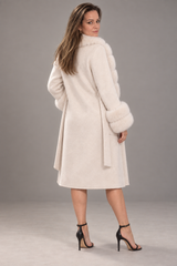 A woman modeling a cream-colored faux fur coat with a high collar, belted waist, and voluminous sleeves, paired with black high-heeled sandals against a neutral gray studio background.