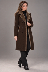 A woman models a long brown double-breasted coat with a brown faux-shearling collar and lining, styled with a black turtleneck, black trousers, and black ankle boots against a solid gray studio background.