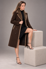 A woman models a long brown double-breasted coat with a brown faux-shearling collar and trim, worn over a black dress and black high-heeled sandals, posing on a light gray two-step stool against a solid gray studio background.