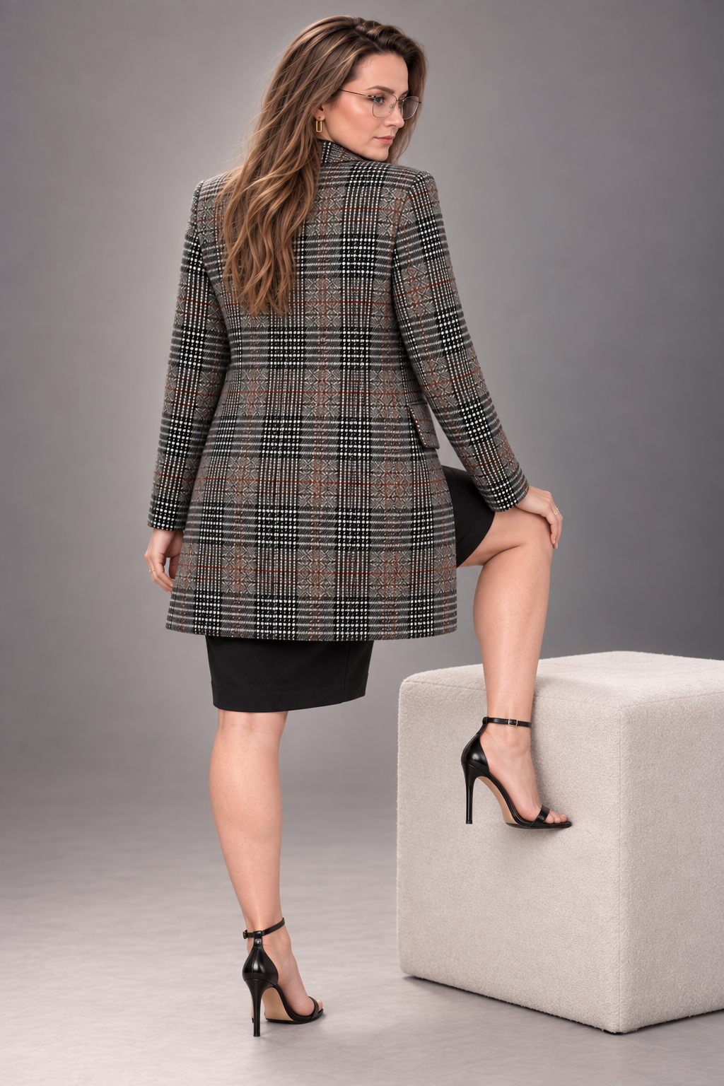 A woman wearing a black, brown, and beige plaid blazer over a black dress, posing next to a light gray cube on a gray studio background.