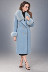 A woman models a light blue double-breasted coat with a fluffy light gray faux fur collar and cuffs, styled with a black top, black skirt, and black high-heeled sandals against a gray studio background.