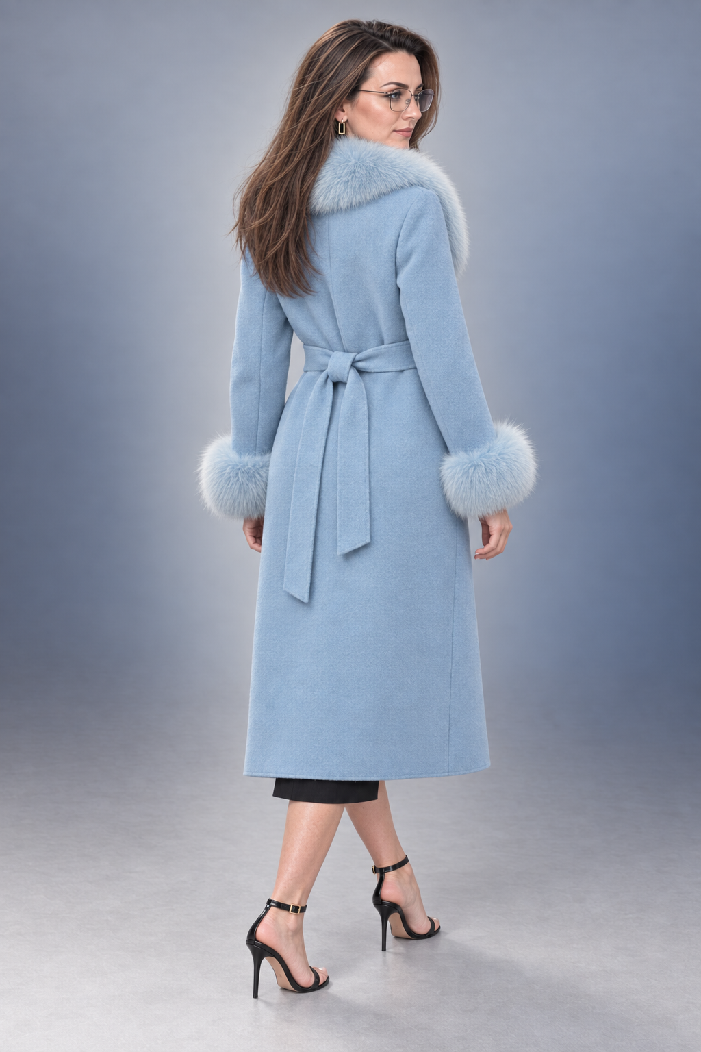 A woman modeling a light blue mid-length coat with a matching waist tie and fluffy light blue fur trim at the collar and cuffs, photographed against a smooth gray studio background.