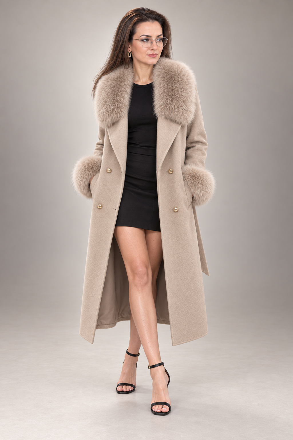 A woman modeling a long beige coat with a large beige faux-fur collar and matching fur-trimmed cuffs, worn over a black mini dress and black strappy heels against a neutral gray studio background.