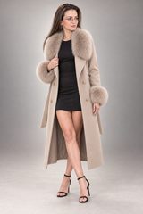 A woman models a long beige coat with a large beige faux-fur collar and cuffs, worn over a black mini dress and black high-heeled sandals, against a neutral gray studio background.