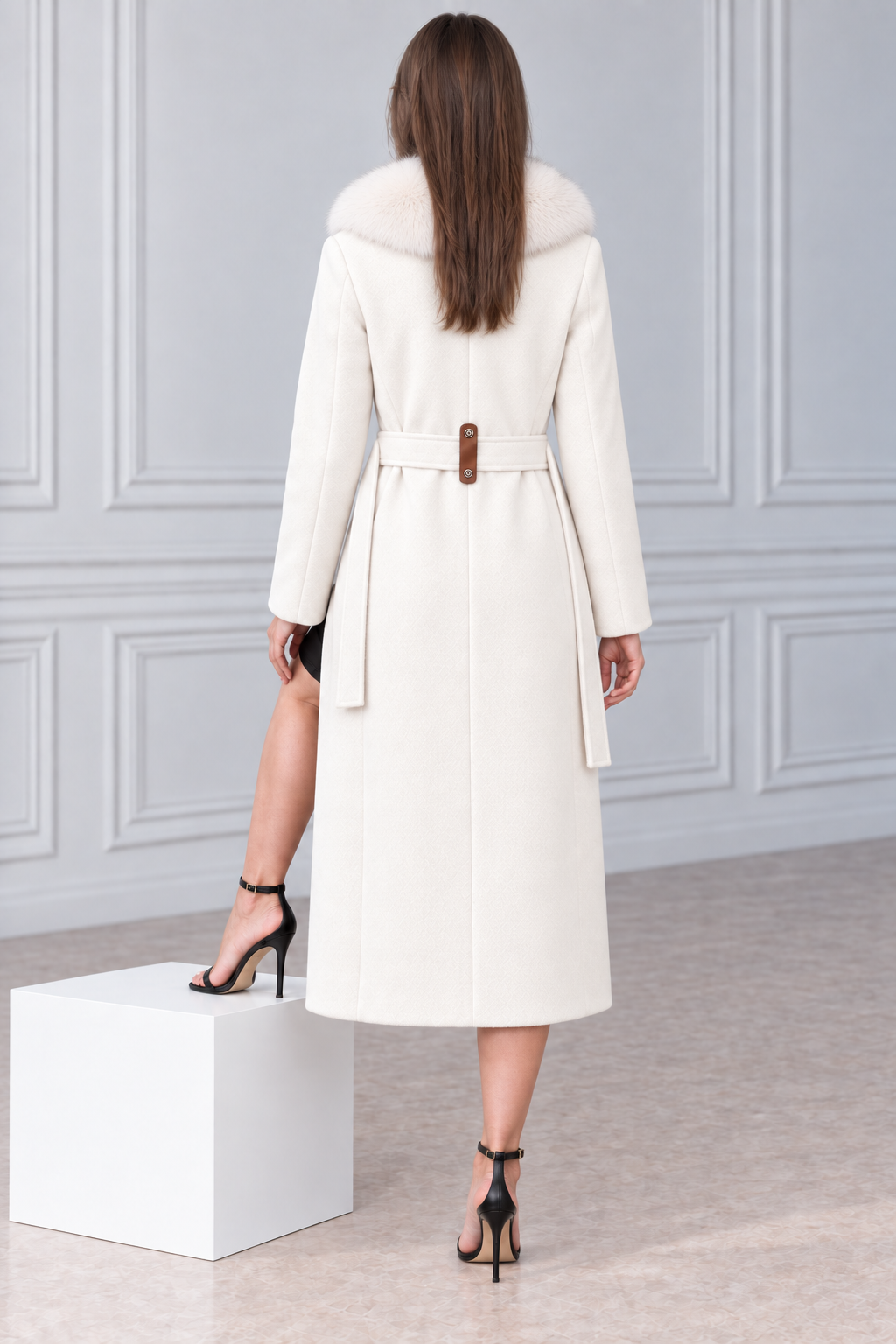 A woman stands with her back to the camera wearing a long cream-colored coat with a fur collar and a brown belt, paired with black high-heeled sandals, in front of a light gray paneled wall.