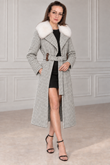 A woman models a long black and white houndstooth coat with a fluffy white fur collar, worn over a black dress and black high-heeled sandals in a light gray paneled room.