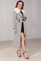 A woman models a long black and white houndstooth coat with a fluffy white fur collar, worn over a black dress and paired with black high-heeled sandals in a classic white paneled room.